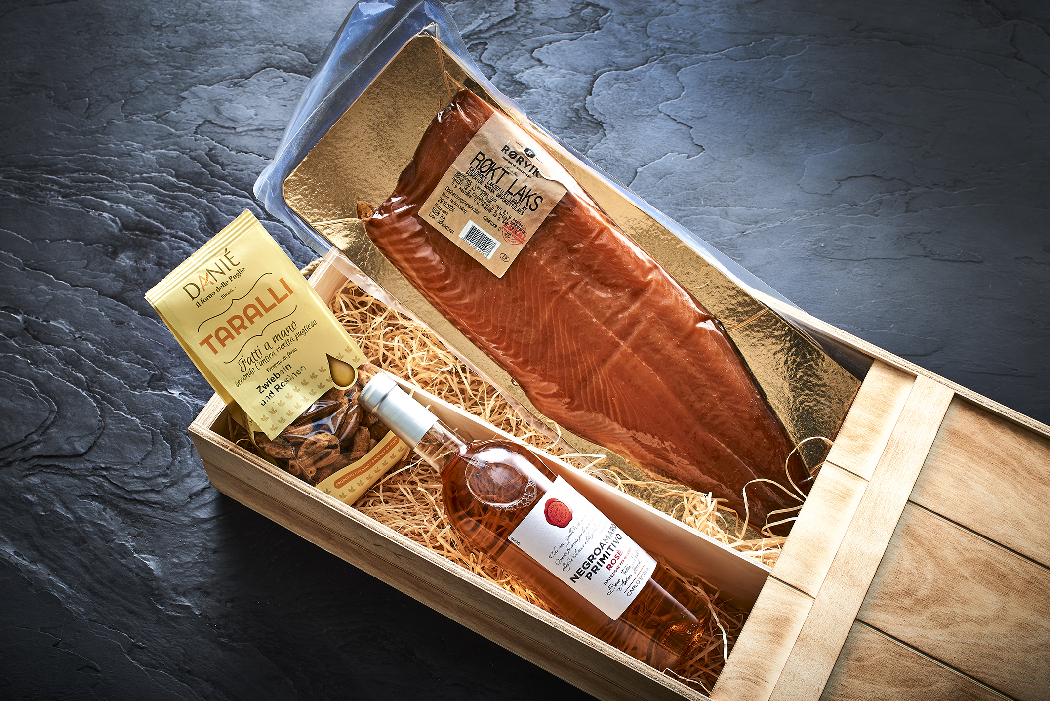 Geschenkbox aus Holz mit geräuchertem Lachs, einer Flasche Roséwein und italienischen Crackern auf einer dunklen Steinoberfläche. – Fishing Adventure GmbH