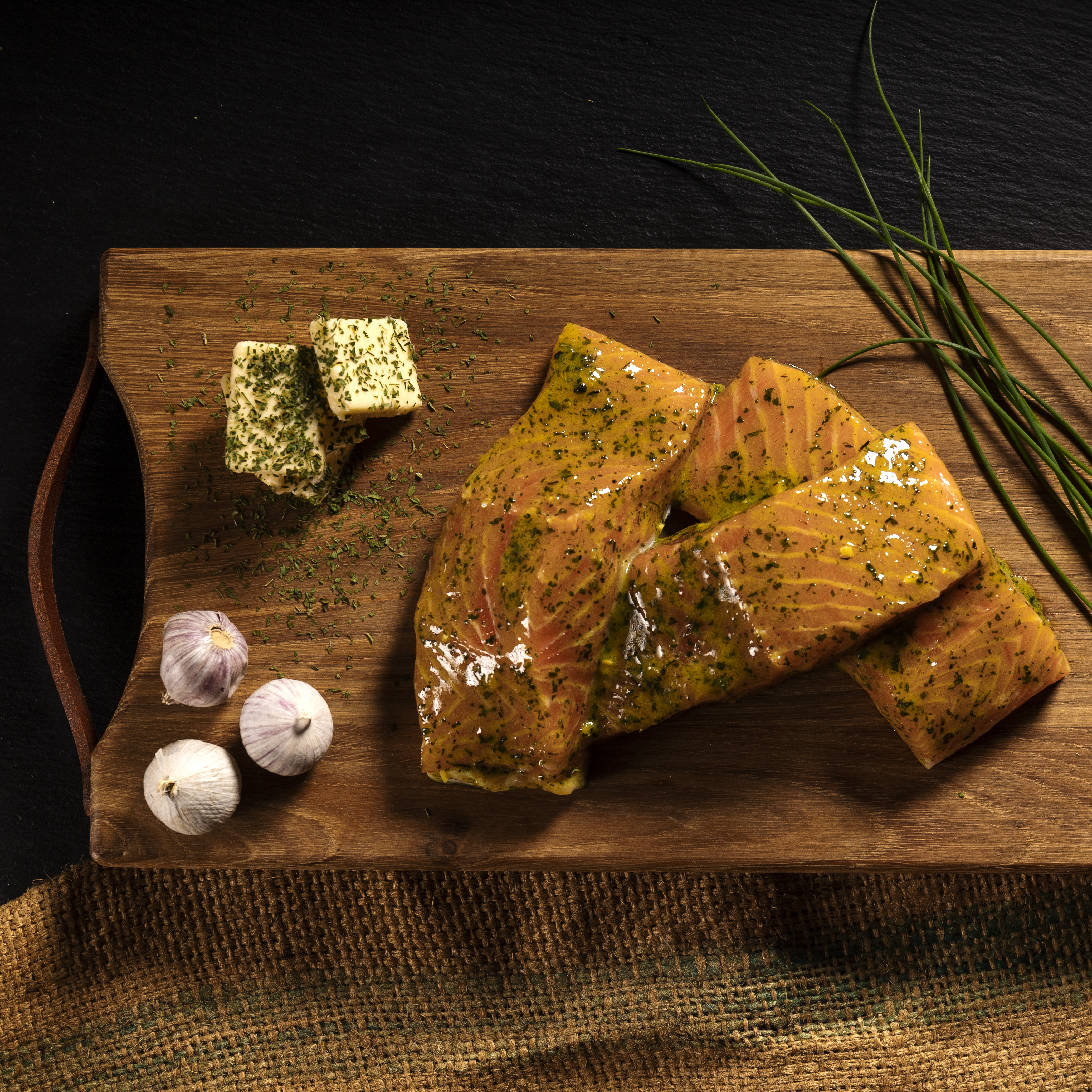 Draufsicht auf gewürzte Lachsfilets auf einem Holzbrett mit Knoblauch und Schnittlauch, bereit zum Kochen. – Fishing Adventure GmbH