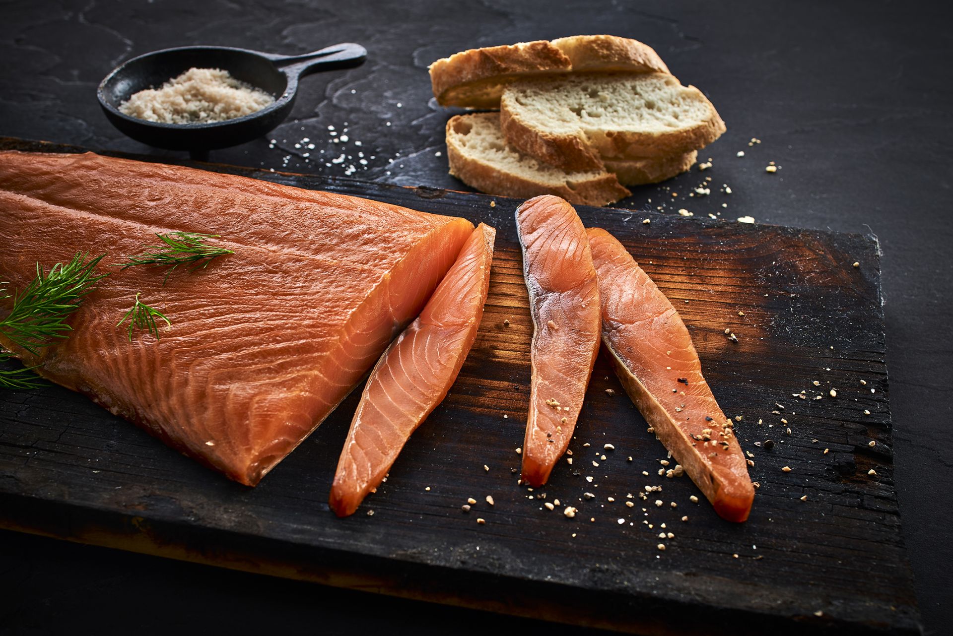 Geräuchertes Lachsfilet mit Scheiben und Brot auf einem dunklen Holzbrett. Salzstreuer im Hintergrund. – Fishing Adventure GmbH