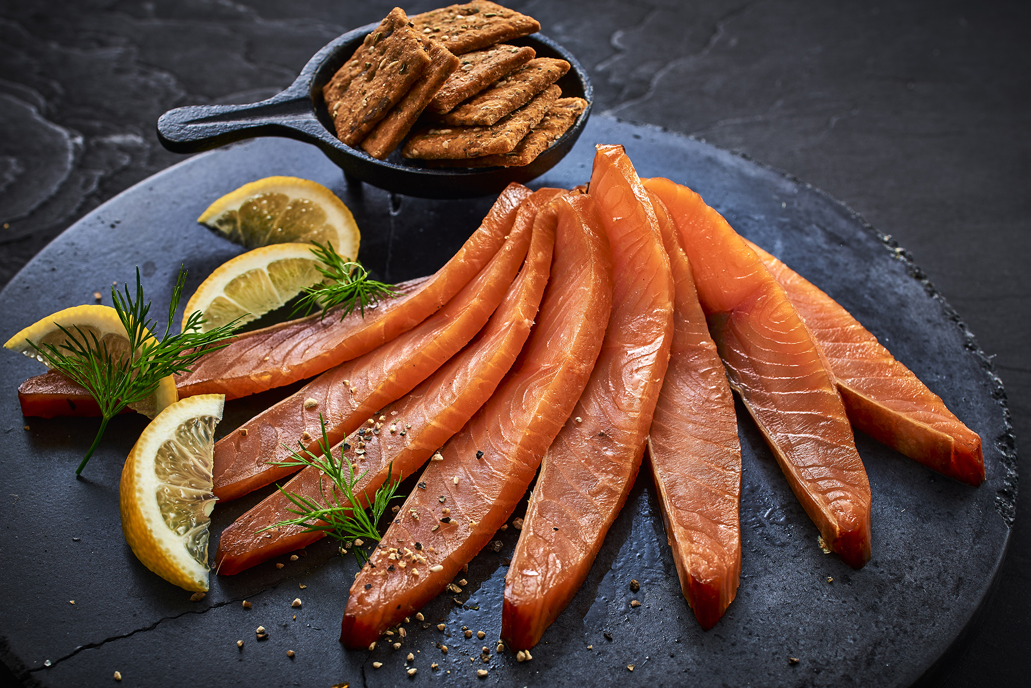 Geräucherte Lachsscheiben, serviert mit Crackern und Zitronenschnitzen auf einer schwarzen Schieferplatte. – Fishing Adventure GmbH