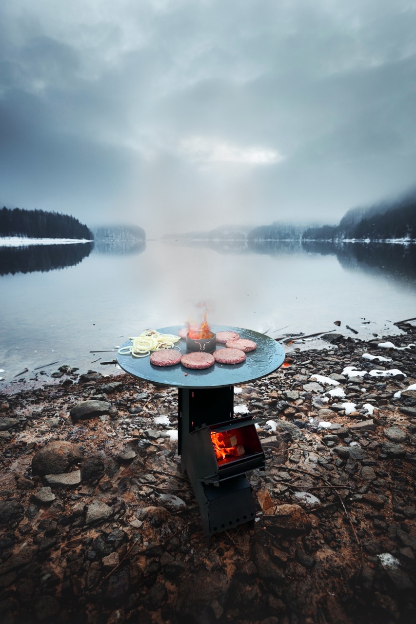 Kochen im Freien auf einem tragbaren Herd neben einem See, mit Burgern und Zwiebeln auf einer Grillplatte. – Fishing Adventure GmbH