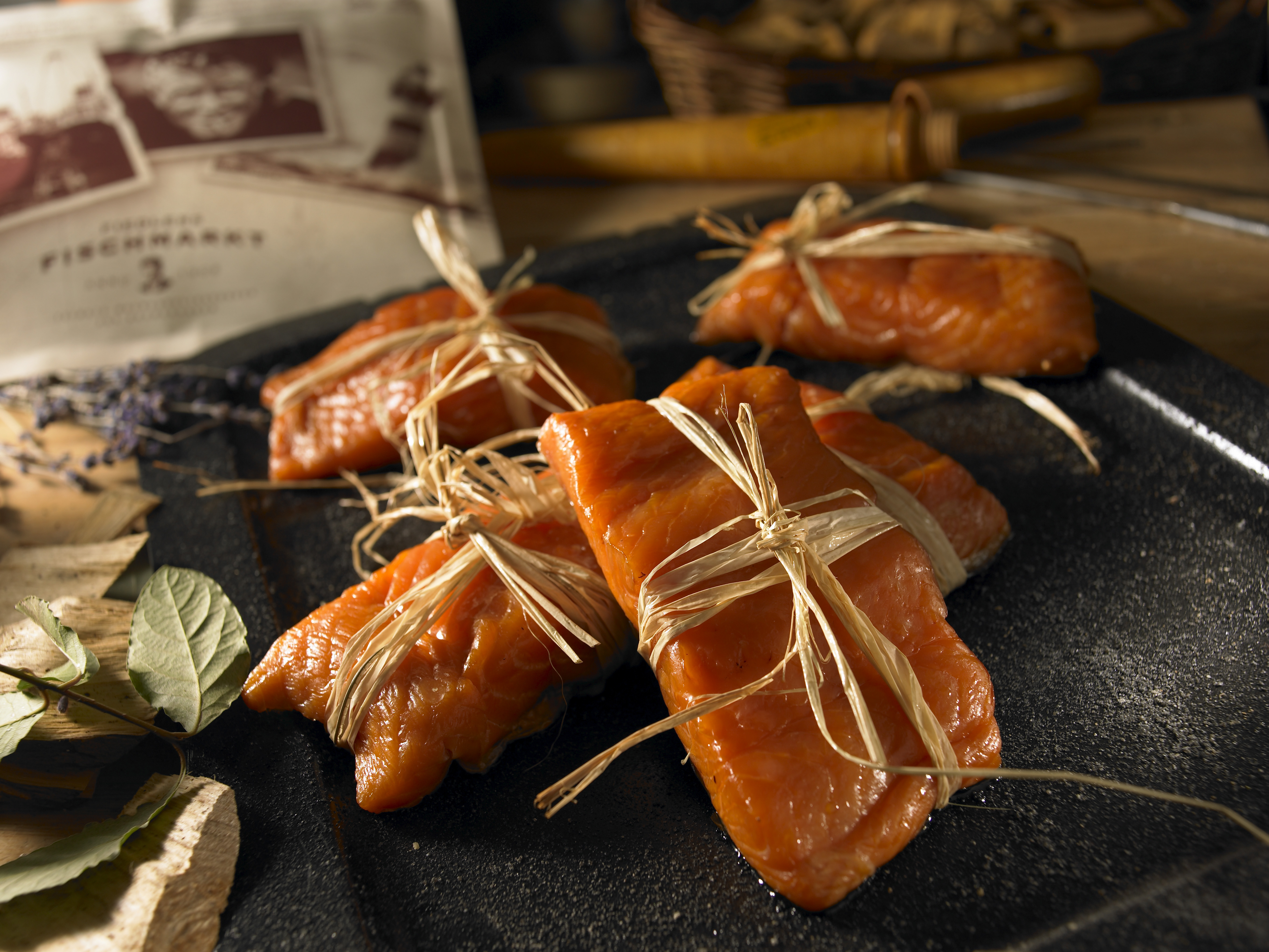 Nahaufnahme von mit Bast zusammengebundenen Räucherlachsfilets auf einem schwarzen Tablett, im Hintergrund Kräuter und Verpackung. – Fishing Adventure GmbH