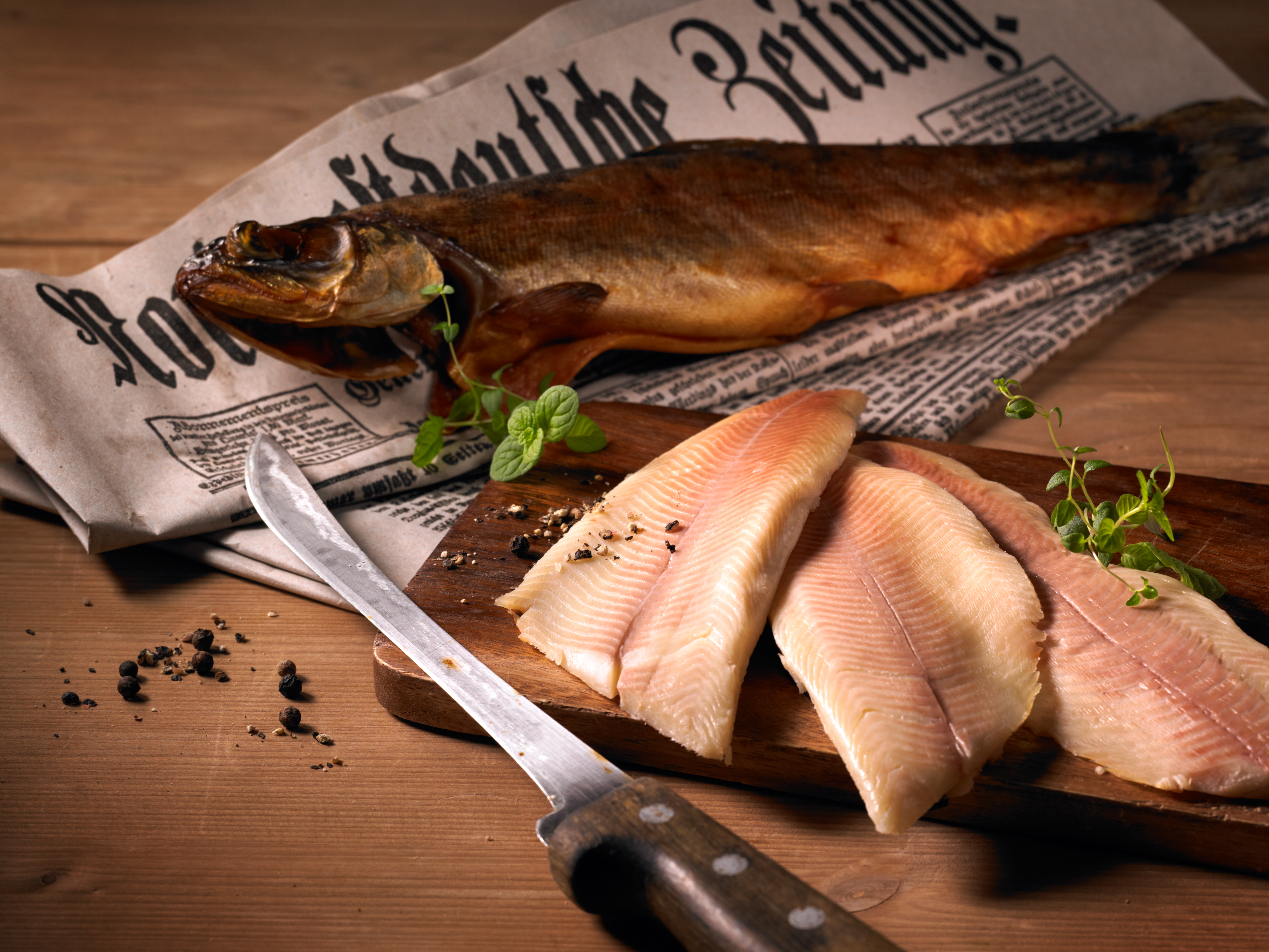 Geräucherte und rohe Fischfilets auf einem Holzbrett mit Messer und Zeitungspapier, Präsentation der Meeresfrüchtezubereitung. – Fishing Adventure GmbH